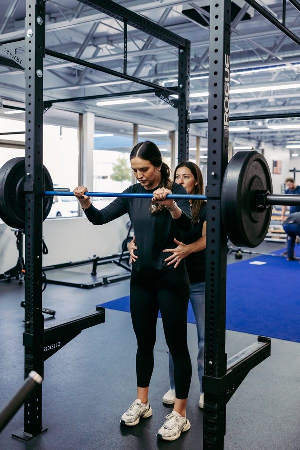 A clinician coaching a woman's barbell squat technique in a squat rack